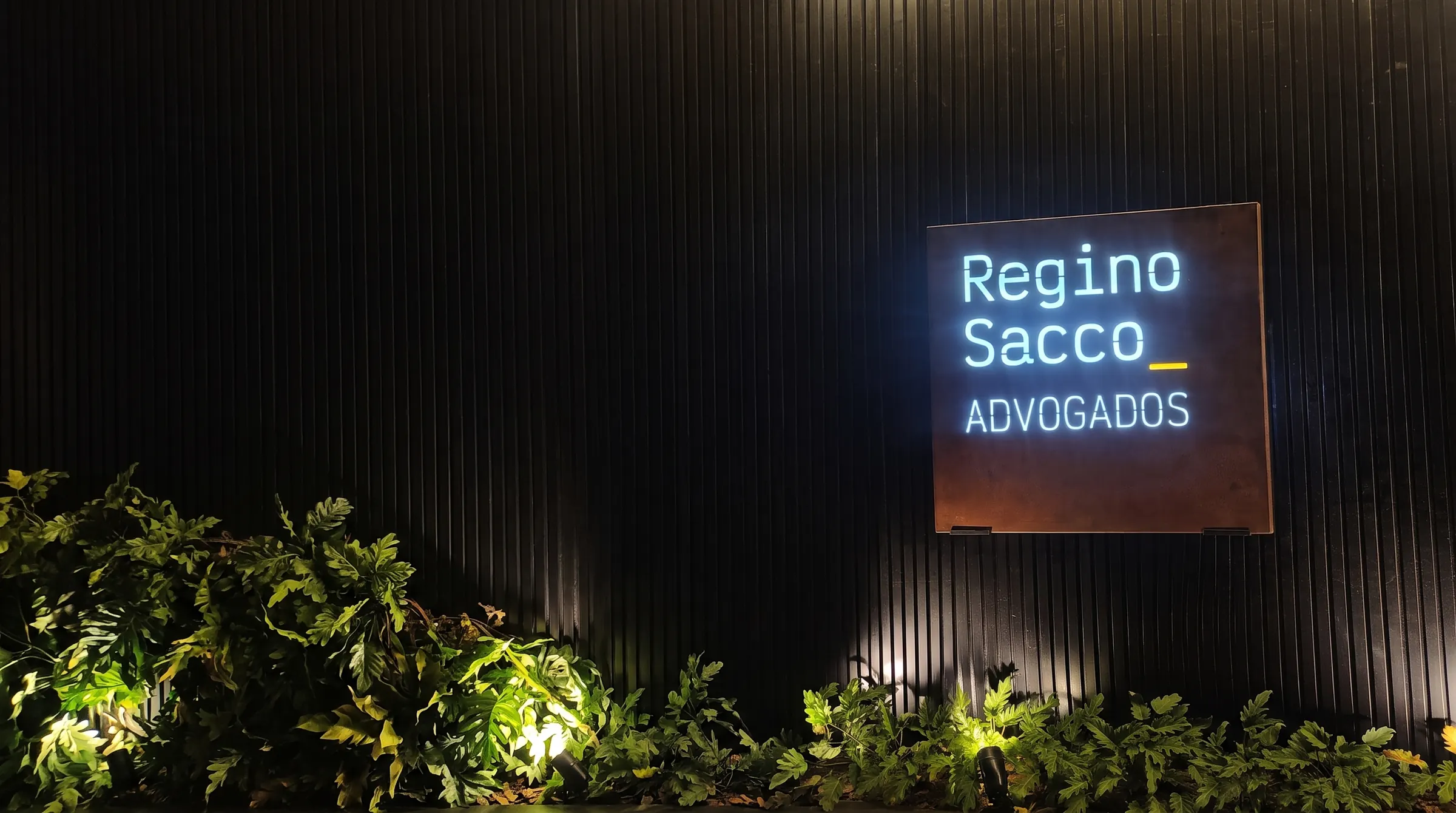 Fachada do escritório RSADV_ — Regino Sacco Advogados, parede ripada com o letreiro iluminado Regino Sacco Advogados
